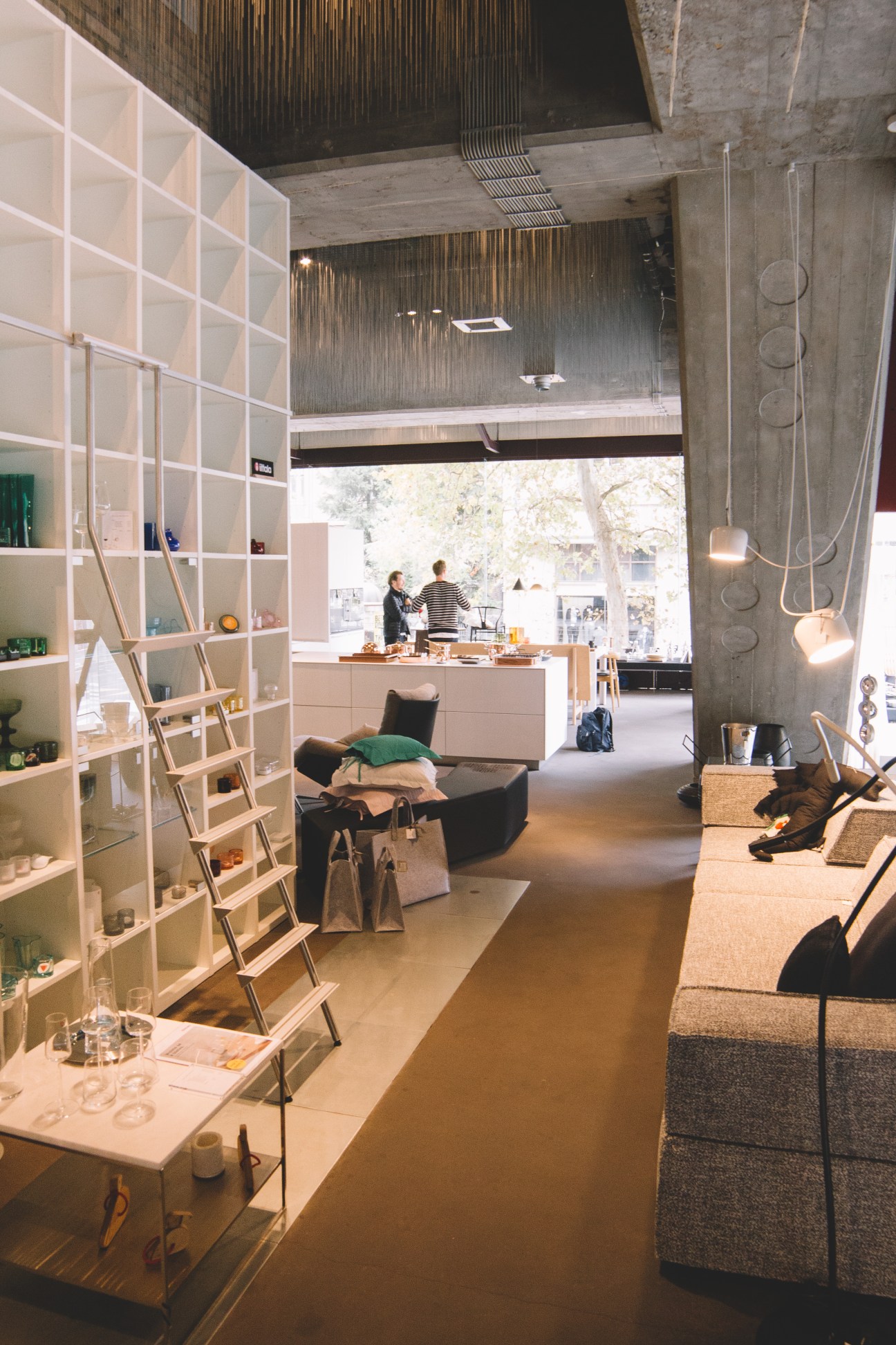 Interior of ID: Doma design shop showing white shelving, concrete columns, pendant lights, and customers browsing furniture.