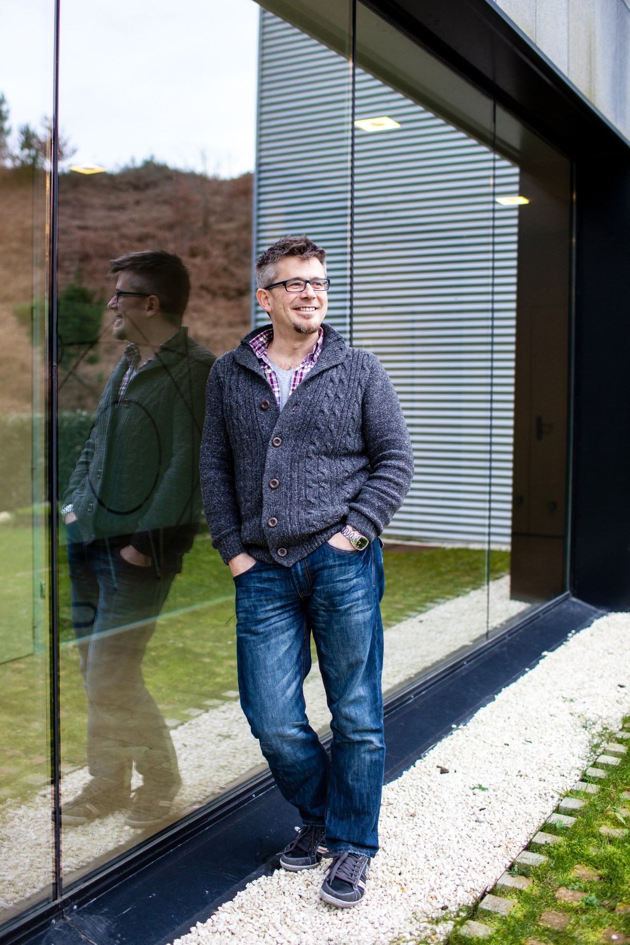Man in glasses and cardigan smiling outside modern building with hillside backdrop