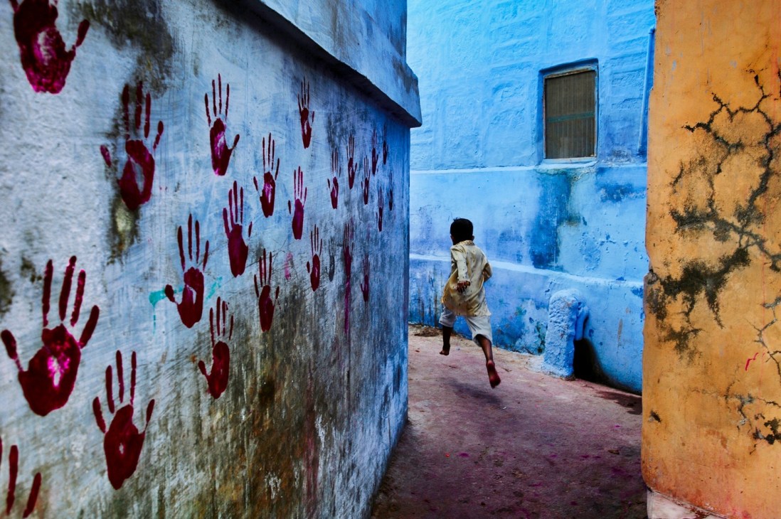 Child running through narrow alleyway with red handprints on white wall and colorful blue and orange buildings.