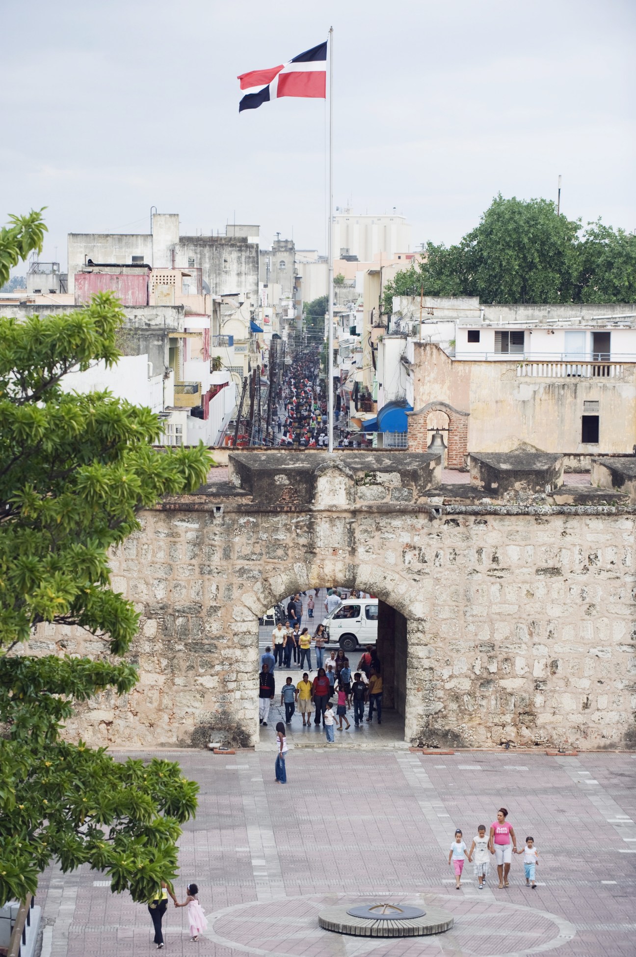 Dominican flag flying above historic stone gates with colonial buildings and crowds in Santo Domingo.