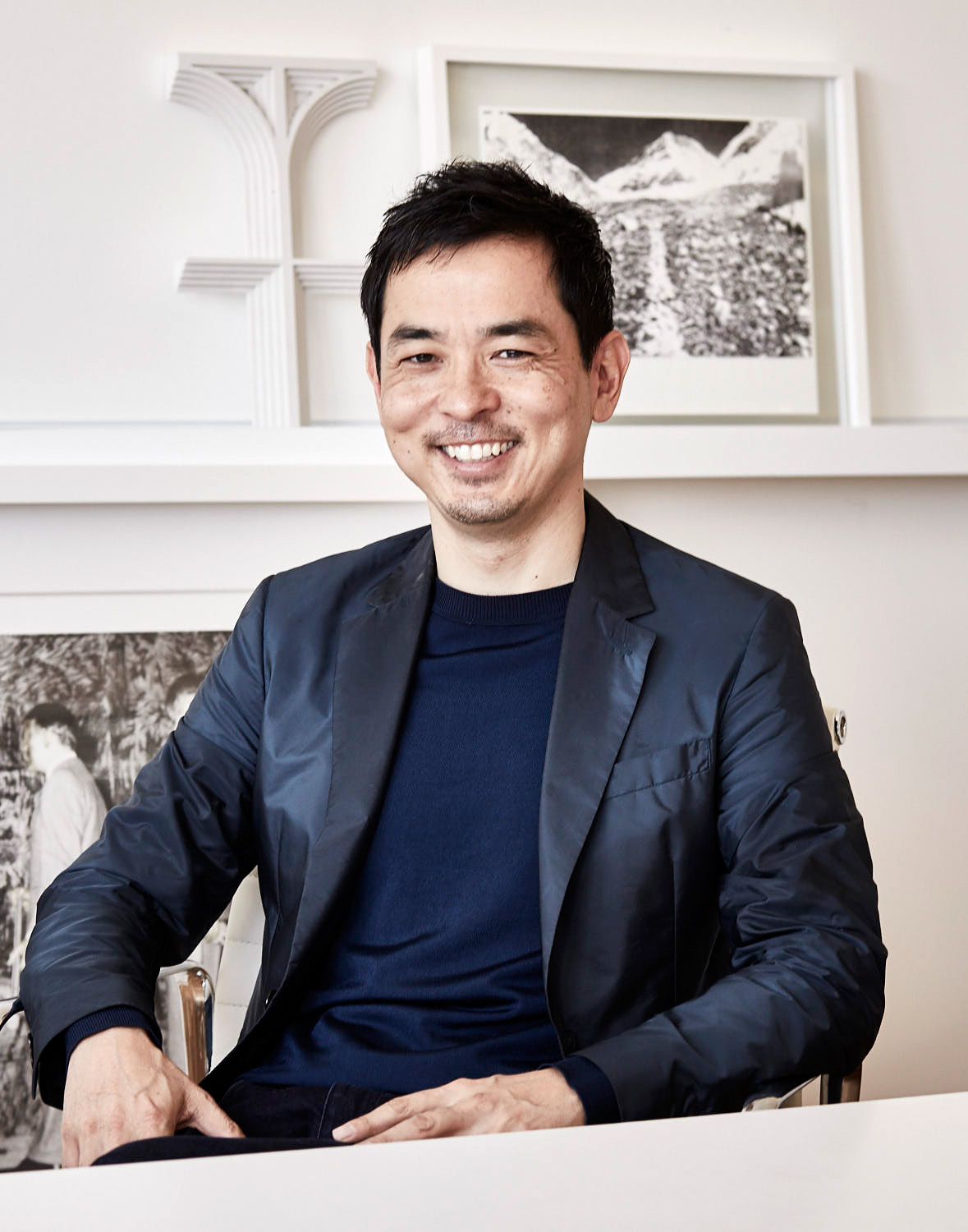 Architect Koichi Takada sitting in a dark blazer and navy sweater, smiling in an interior with white shelving