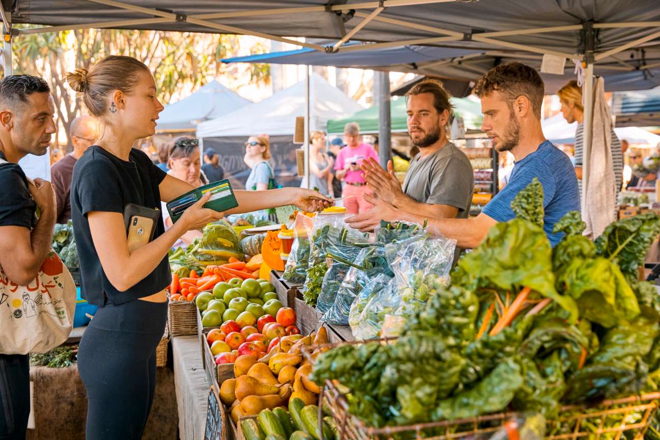 A woman in a black shirt makes a payment at an outdoor farmers market produce stall displaying fresh organic vegetables.