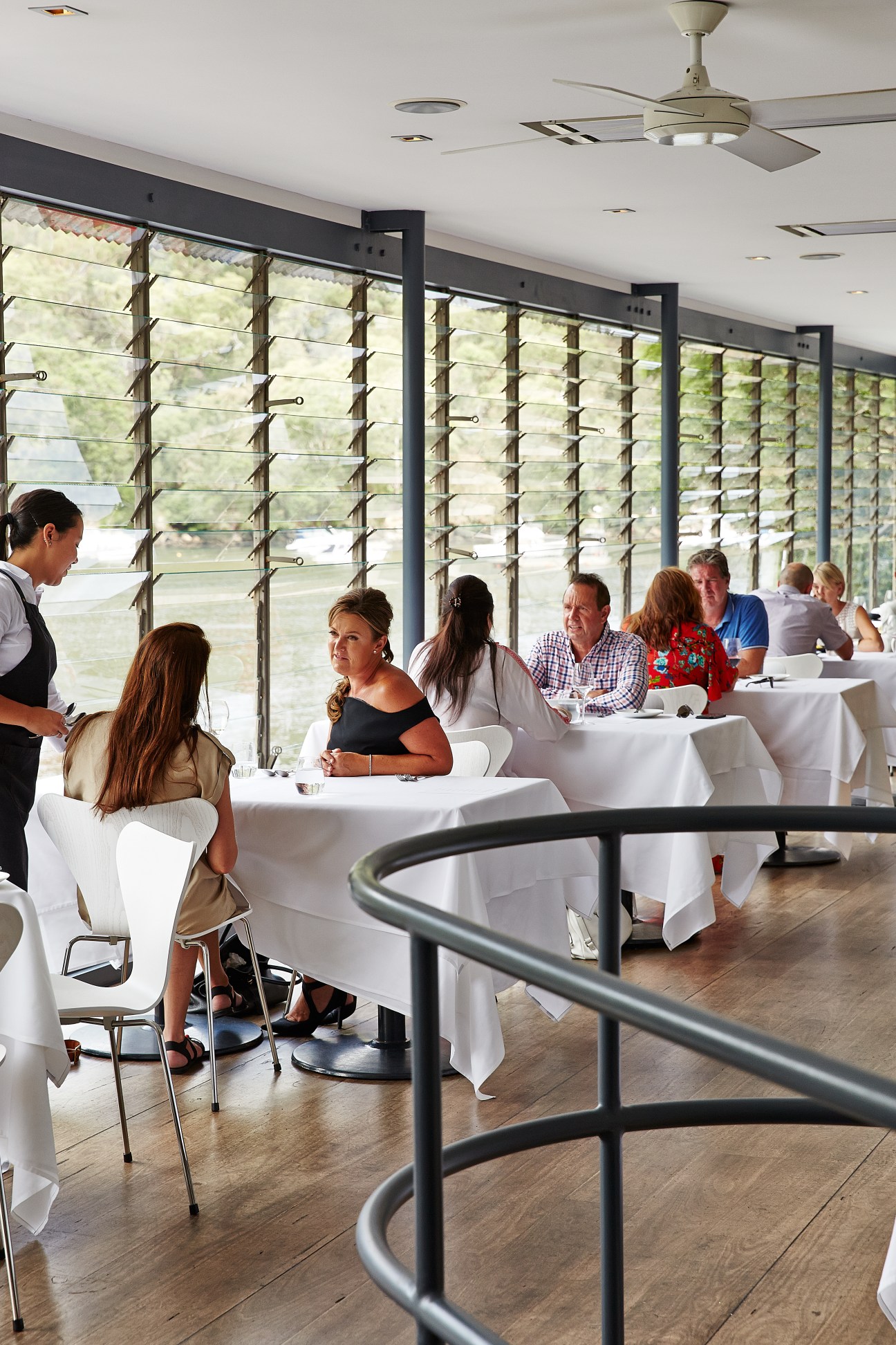 Diners seated at white-clothed tables in a bright restaurant with louvered windows overlooking a river and bush landscape.