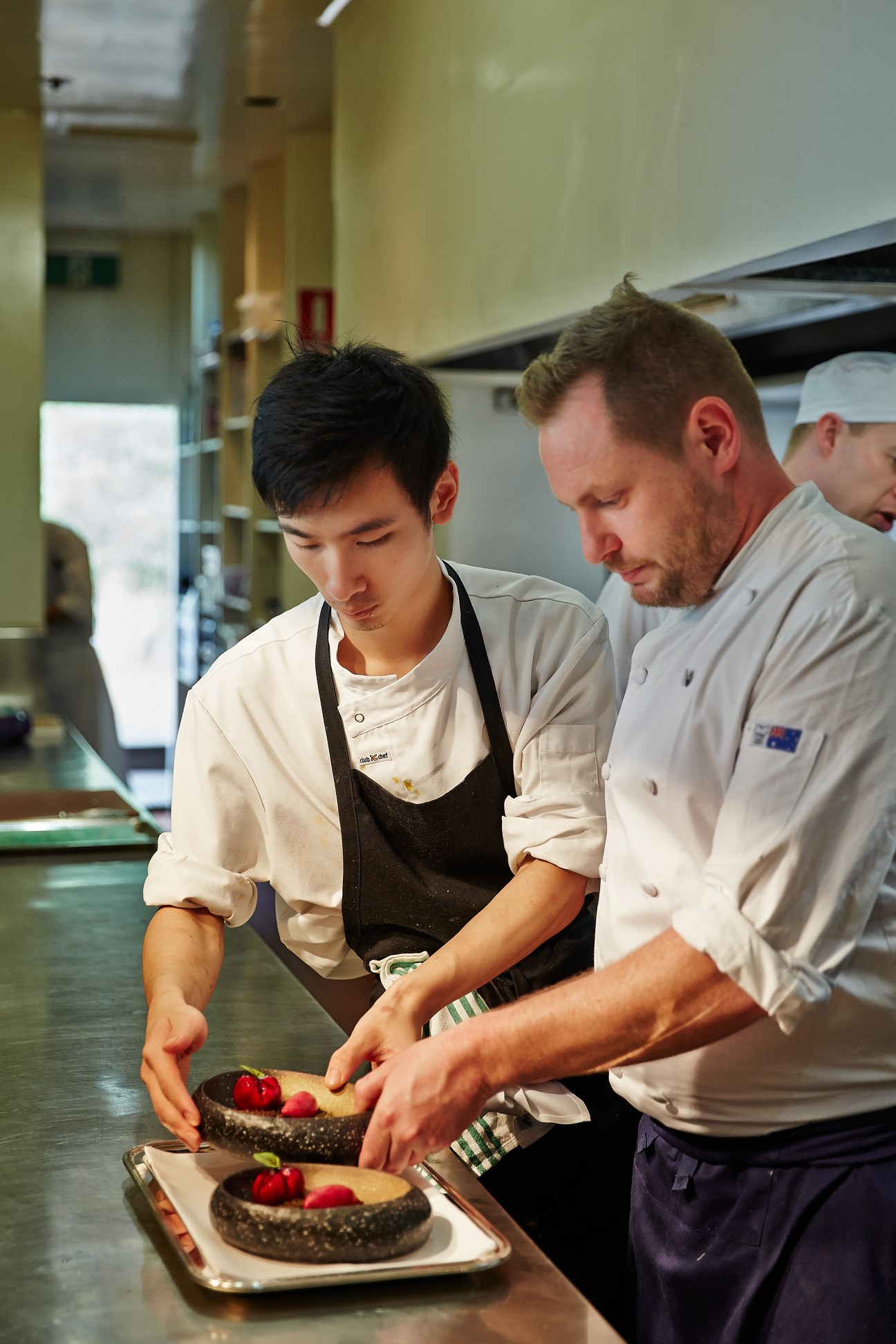 Two chefs plate a dark dessert with red berries in a professional kitchen.