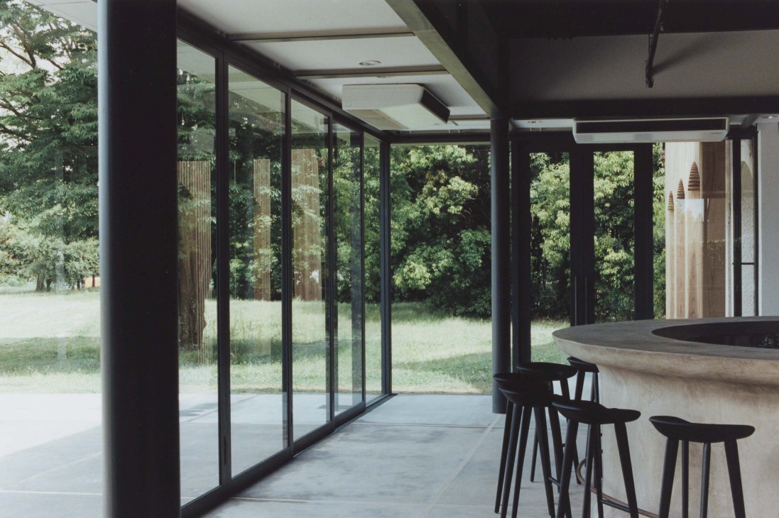 Modern interior with black-framed glass doors opening to forest views, concrete flooring, and wooden bar counter with black stools.