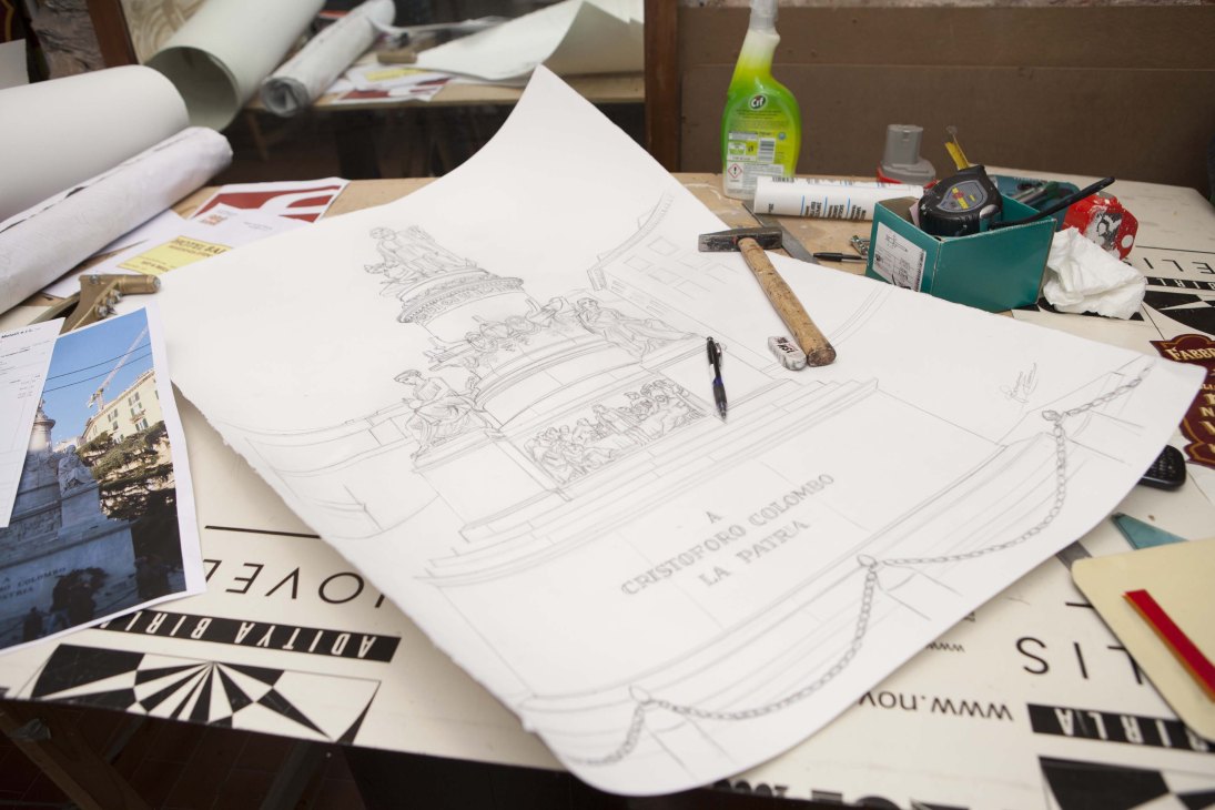 Hand-drawn pencil sketch of ornate lettering design on paper at a workbench with tools and reference photos.