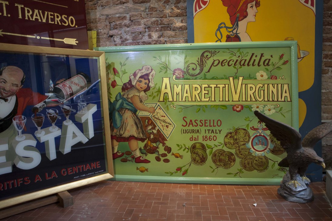 Vintage Italian product advertisements displayed in a workshop, including Amaretti Virginia and other colorful tin signs with ornate typography.