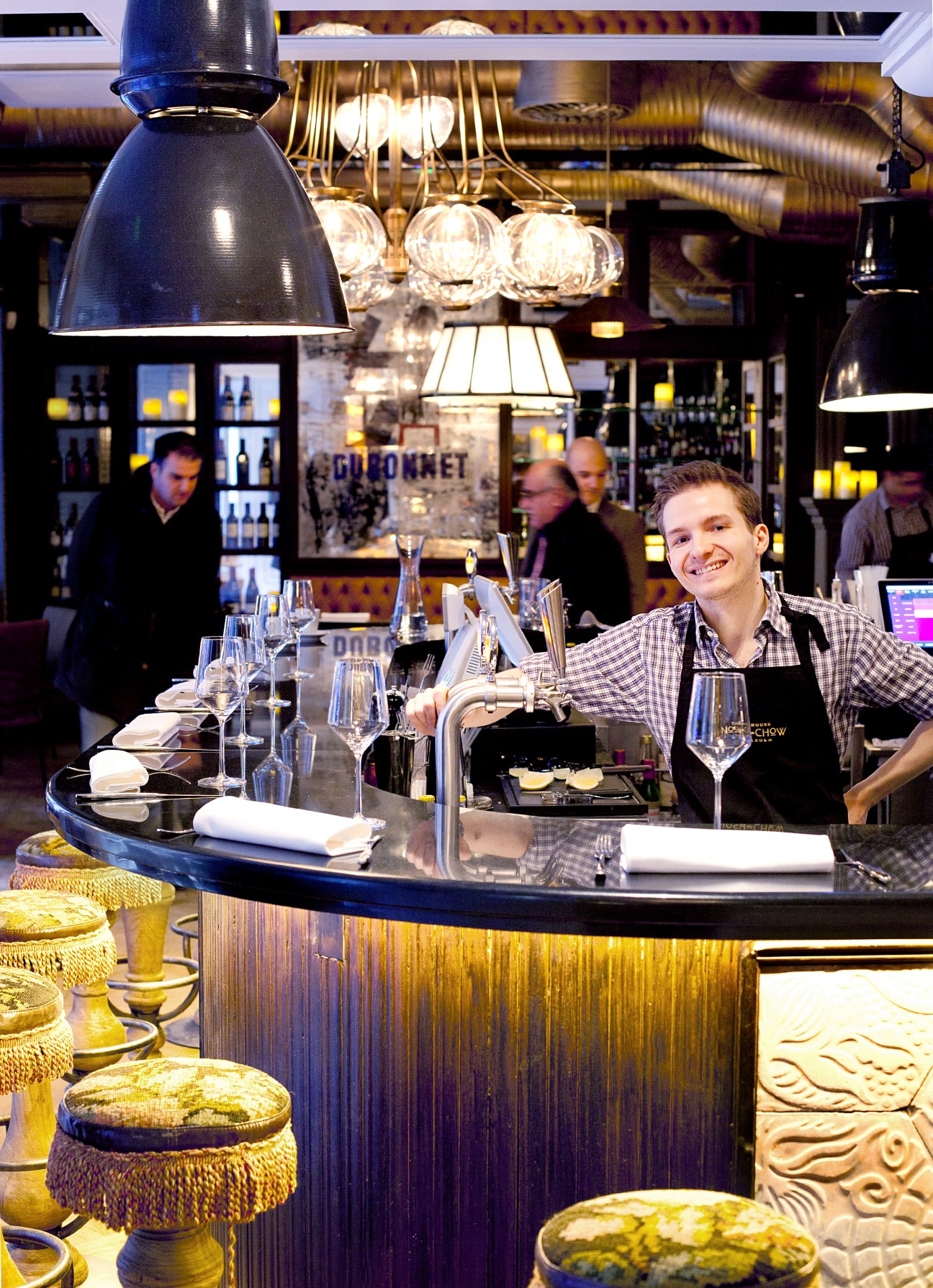 Smiling bartender stands behind a modern bar counter with hanging pendant lights at Nosh and Chow restaurant in Stockholm.