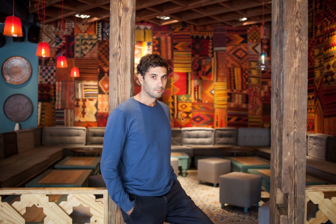 Man in blue sweater standing in modern hotel lounge with colorful patterned rugs and red pendant lights.