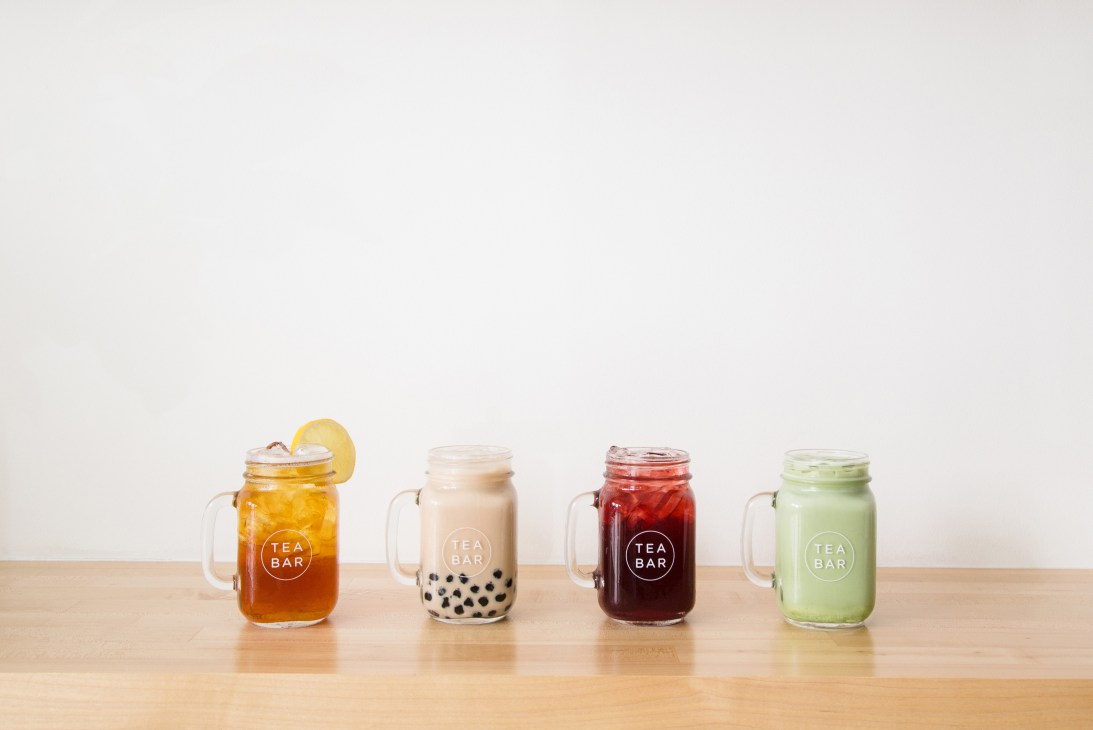 Four mason jars with handles containing different colored beverages, each labeled 'TEA BAR' on a wooden counter.