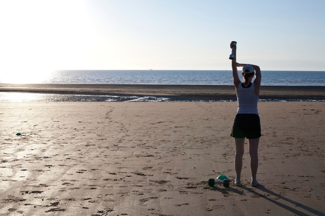 Woman exercising with dumbbells on a sandy beach with ocean in the background.