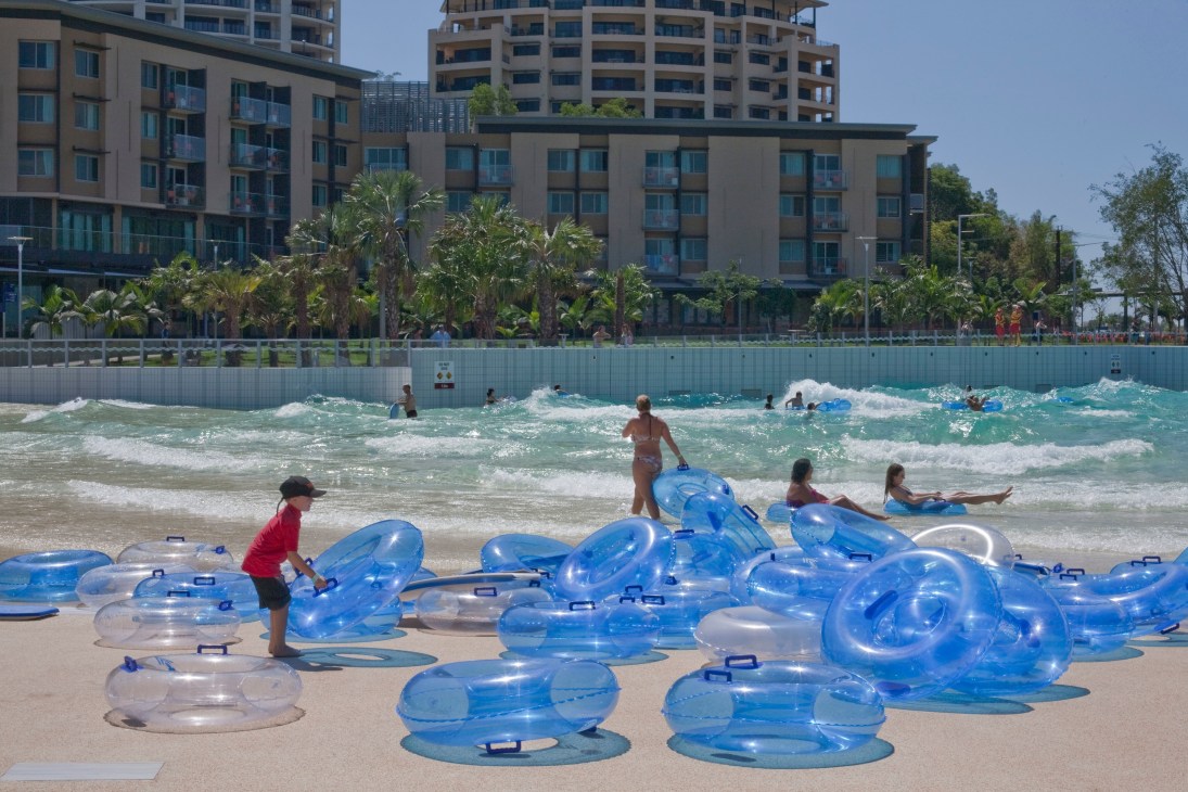 Beachgoers enjoying a wave pool with blue inflatable floats near modern residential buildings.