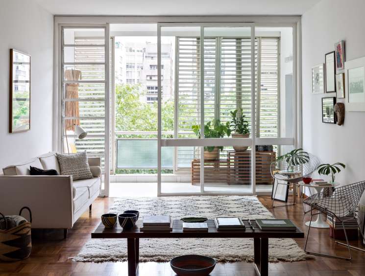 Modernist living room with large glass doors opening to a balcony with horizontal shutters, neutral sofa, dark coffee table, potted plants, and framed artwork on walls.
