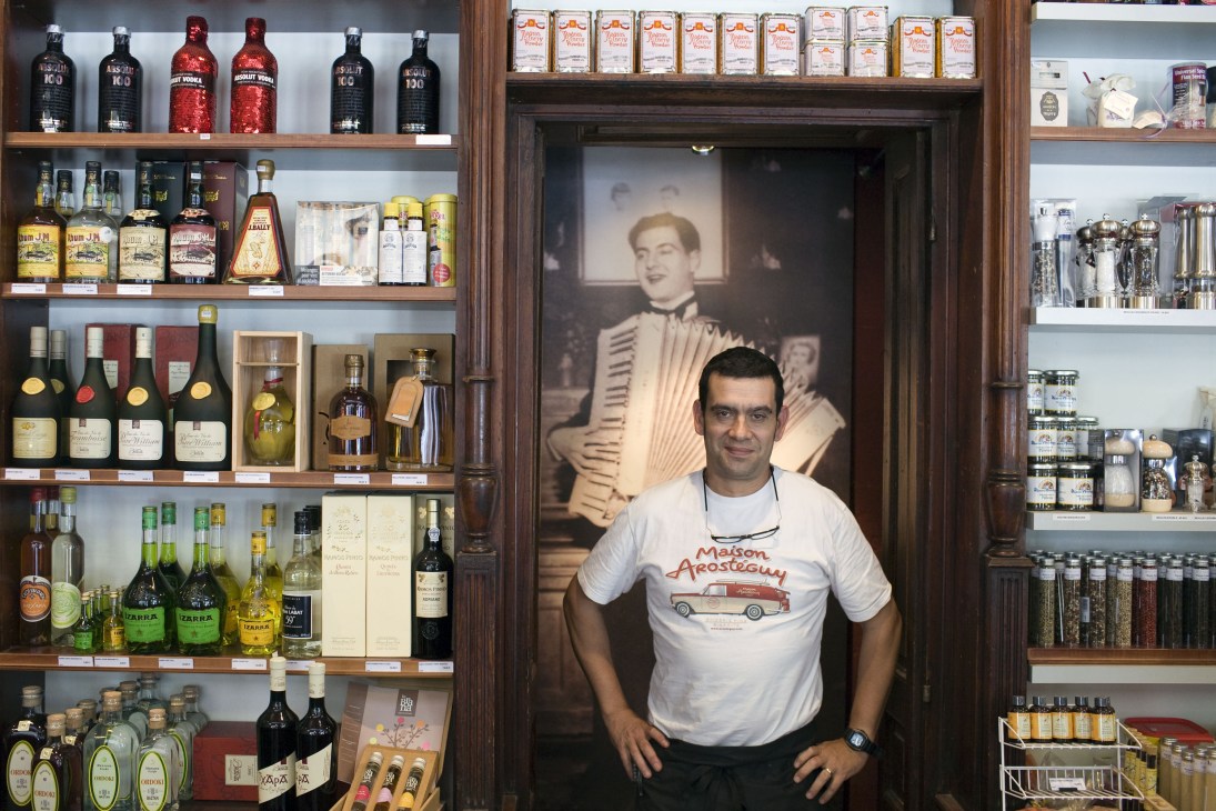 Pierre Arostéguy stands in front of vintage photograph at Maison Arostéguy, surrounded by wine bottles and gourmet products on shelves.