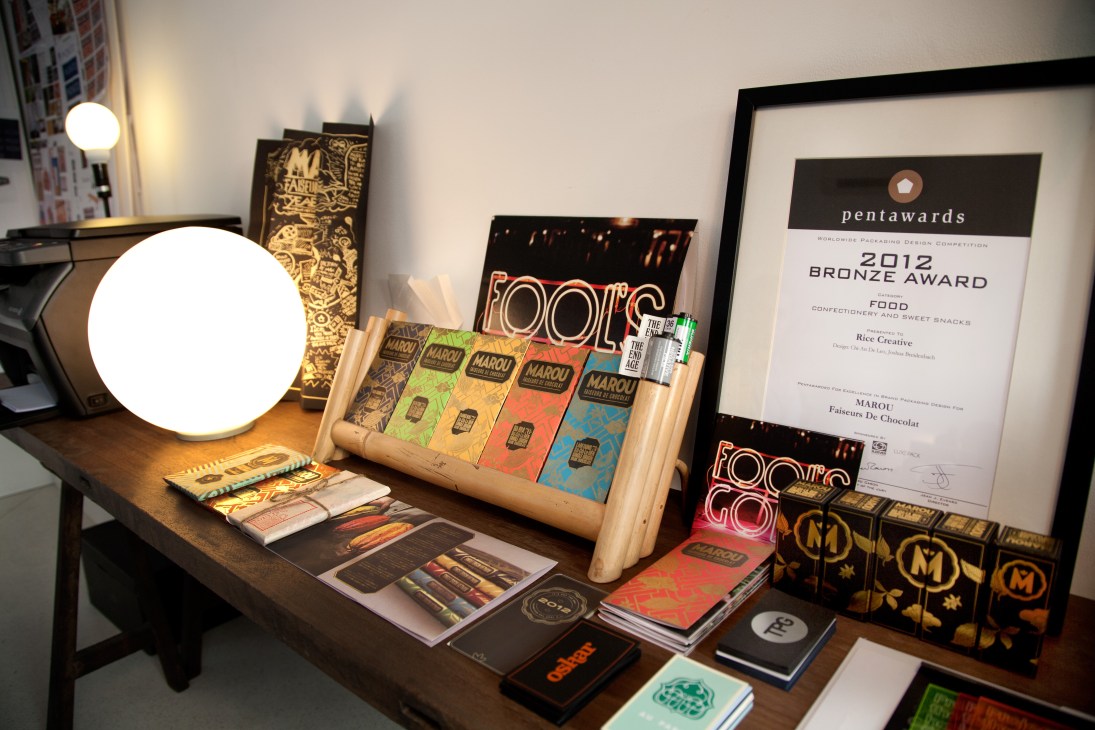 Rice Creative office display with Marou chocolate packaging, design awards, and printed materials on wooden desk.