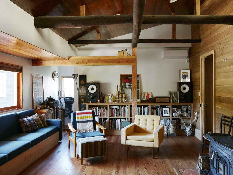 Living room with exposed wooden beams, handmade furniture including blue sofa and cream armchair, bookshelves, and natural lighting.