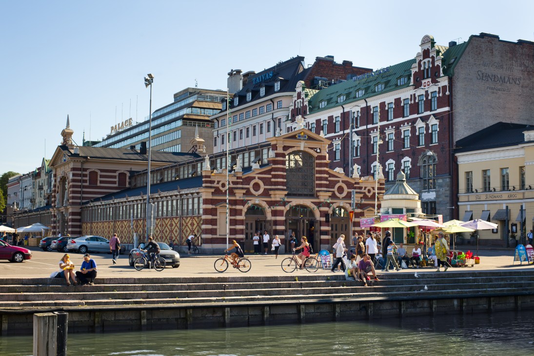 Neo-renaissance Vanha Kauppahalli market building with red and cream striped facade on Helsinki waterfront, surrounded by modern buildings and people by the harbor.