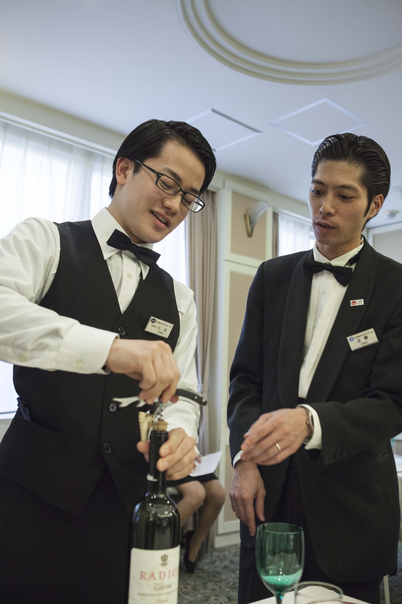 Instructor Shinji Ueno demonstrates wine bottle opening technique to a student in formal service attire.