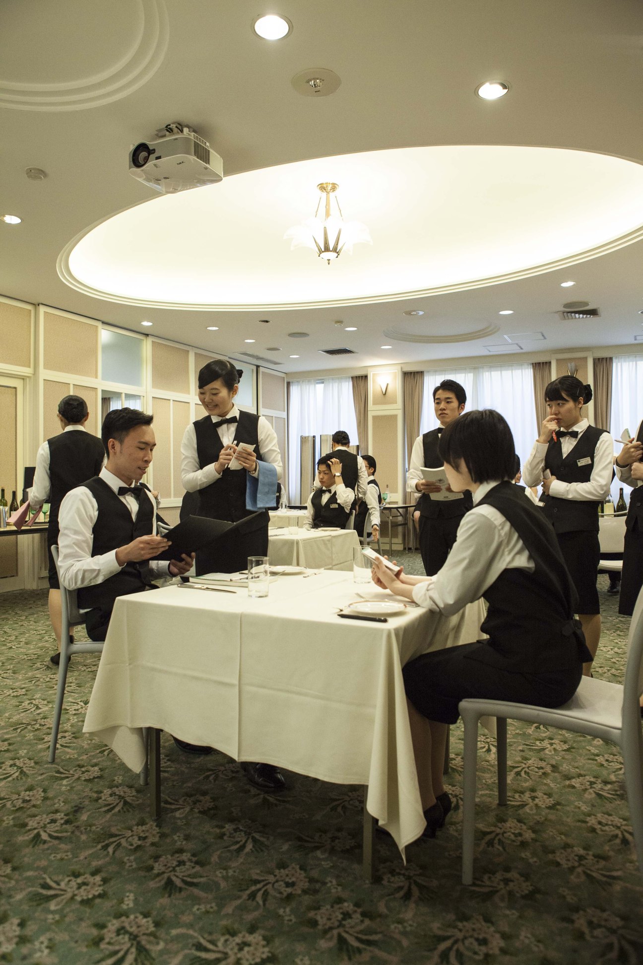 Japan Hotel School students in formal waitstaff uniforms role-playing a restaurant service in a training classroom.