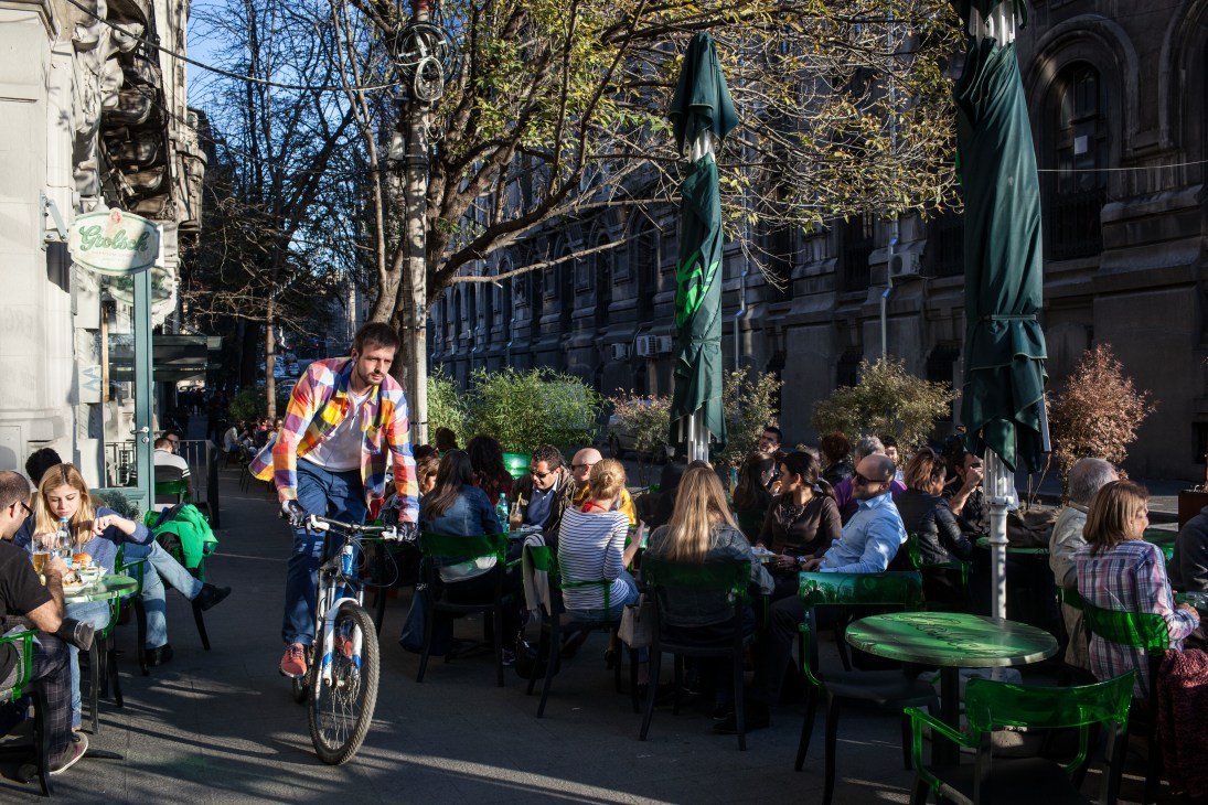 Outdoor café scene on Lipscani Street with people seated at green tables and a man on a bicycle.