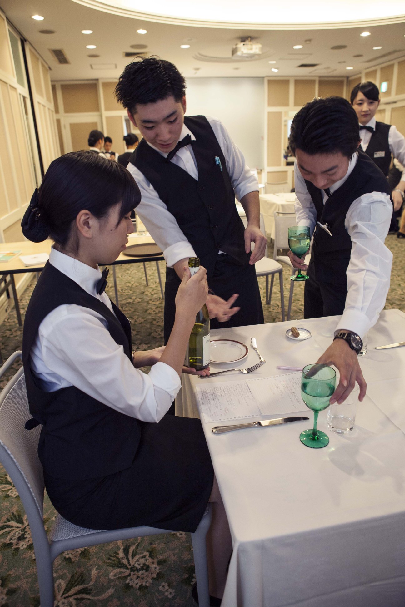 Japan Hotel School students in formal service uniforms practicing wine service at a table.