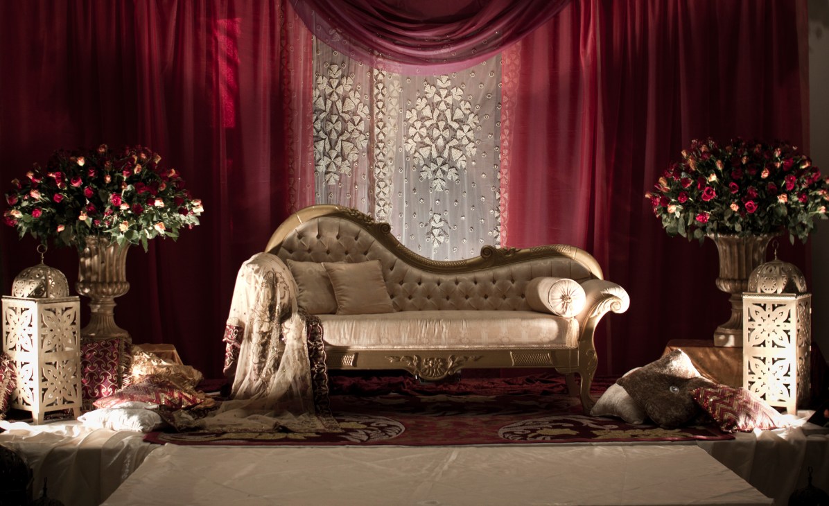 Ornate gold sofa with cushioned seat under burgundy draped fabric, flanked by floral arrangements in Corinthia Hotel wedding hall.