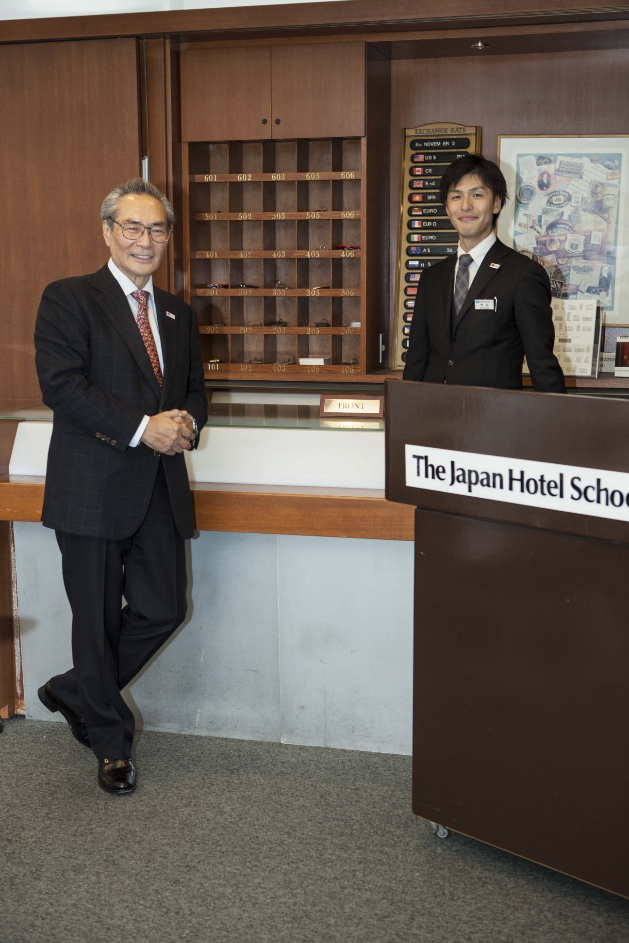 President Tsutomo Ishizuka stands at a reception desk in the Japan Hotel School lobby with a world currency exchange display behind him.