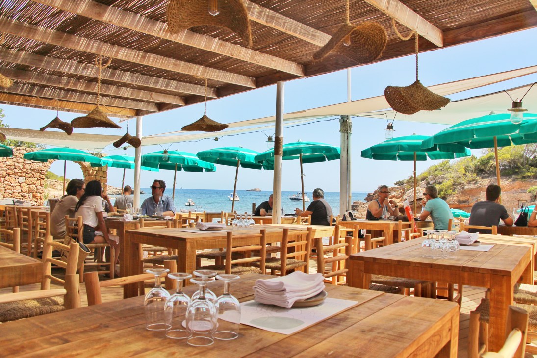 Beachside Mediterranean restaurant Cala Bonita with turquoise umbrellas, wooden furniture and sea views in Ibiza.