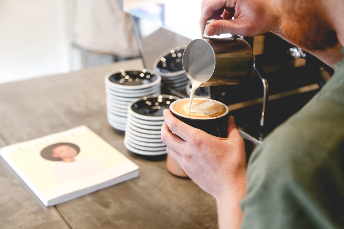 A barista pours steamed milk into a black cup of espresso, creating latte art.