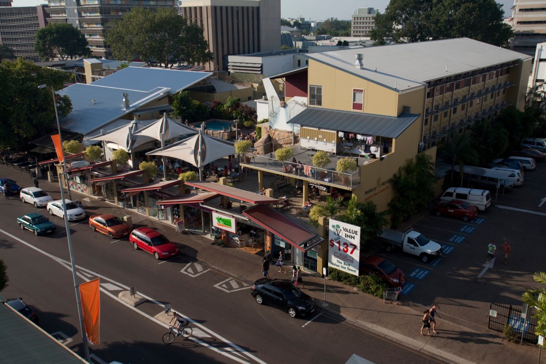 Aerial view of Melaleuca On Mitchell hostel with yellow building, red metal structures, and street-level shops in Darwin.