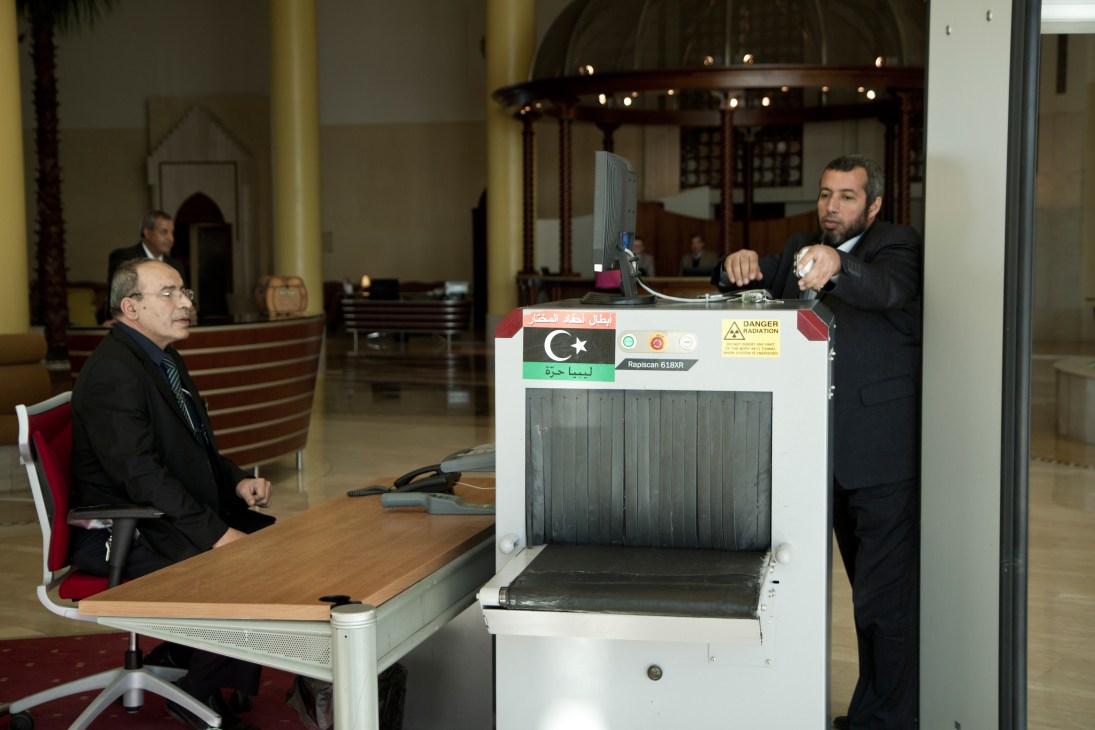 Man operating a metal detector screening device in a hotel lobby with Libyan flag imagery.