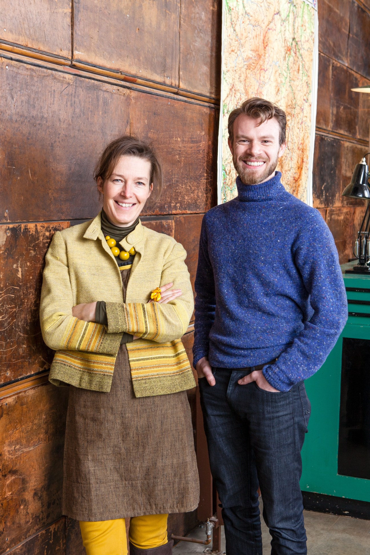 Maria Speake in a yellow jacket stands next to James Stevens in a blue sweater against a wooden wall.