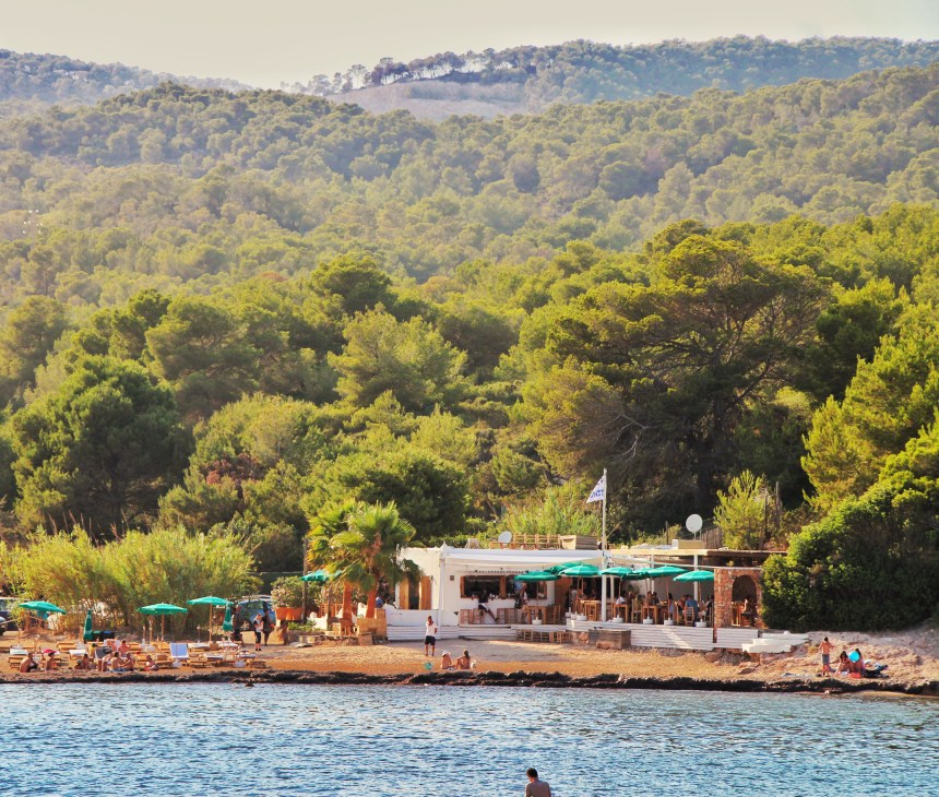 Cala Bonita beach restaurant with turquoise umbrellas on Ibiza shoreline backed by pine forest.