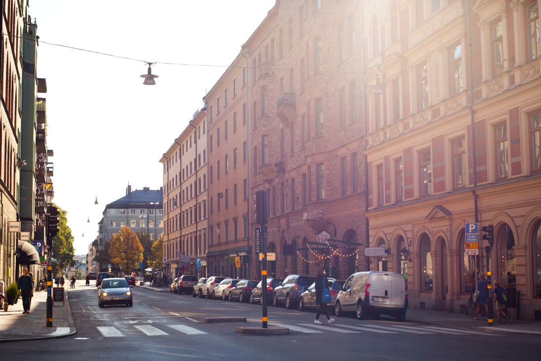 A Stockholm street lined with red-brick historic buildings, parked cars, pedestrians, and street lamps on an autumn day.