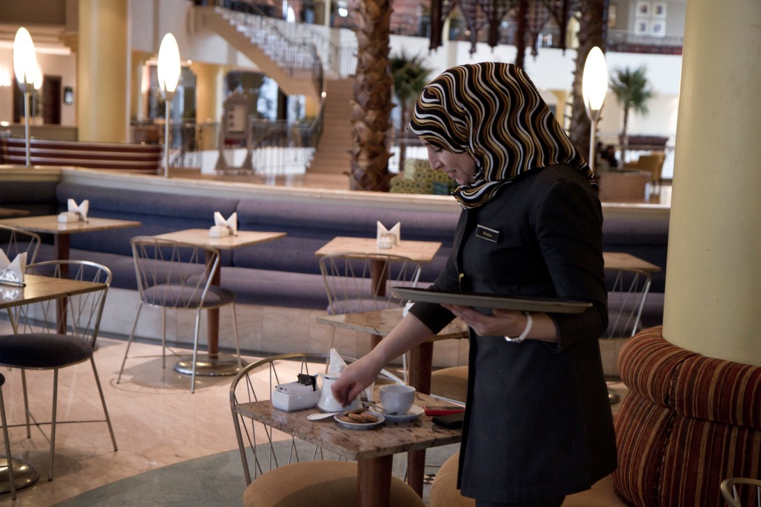 Hotel staff member Malika serves coffee and cookies to guests in a modern lobby café.