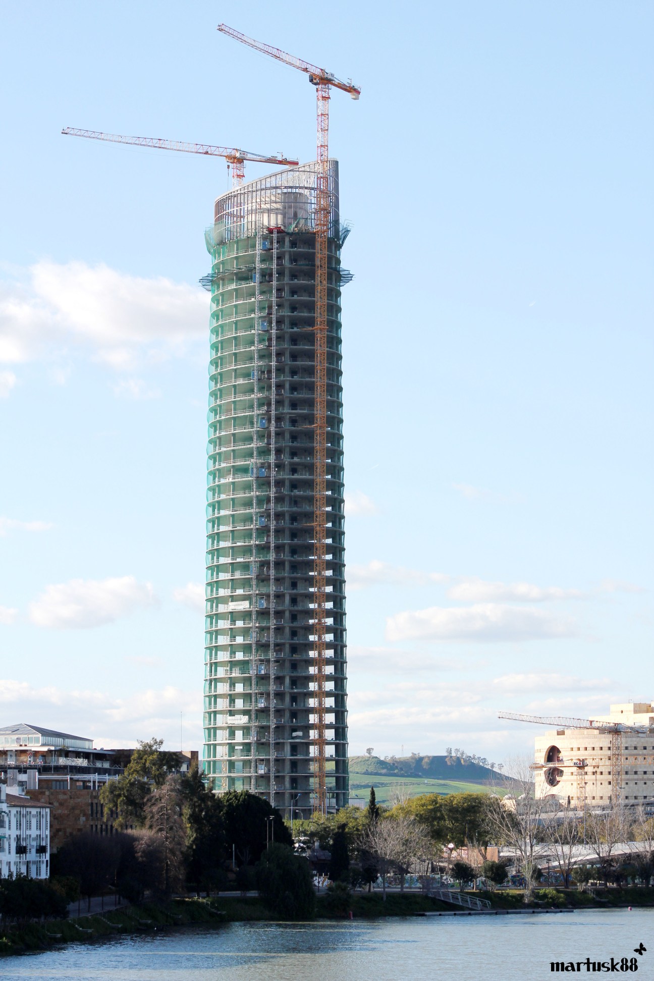 Torre Pelli, an unfinished green-glassed skyscraper under construction with cranes, rising above Seville's riverside.