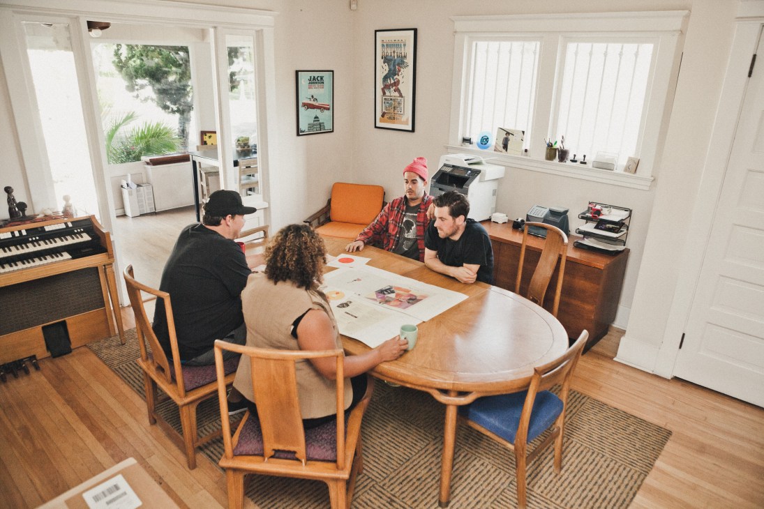 Four people discussing artwork around a wooden dining table at Brushfire Records office with large windows and vintage synthesizer.