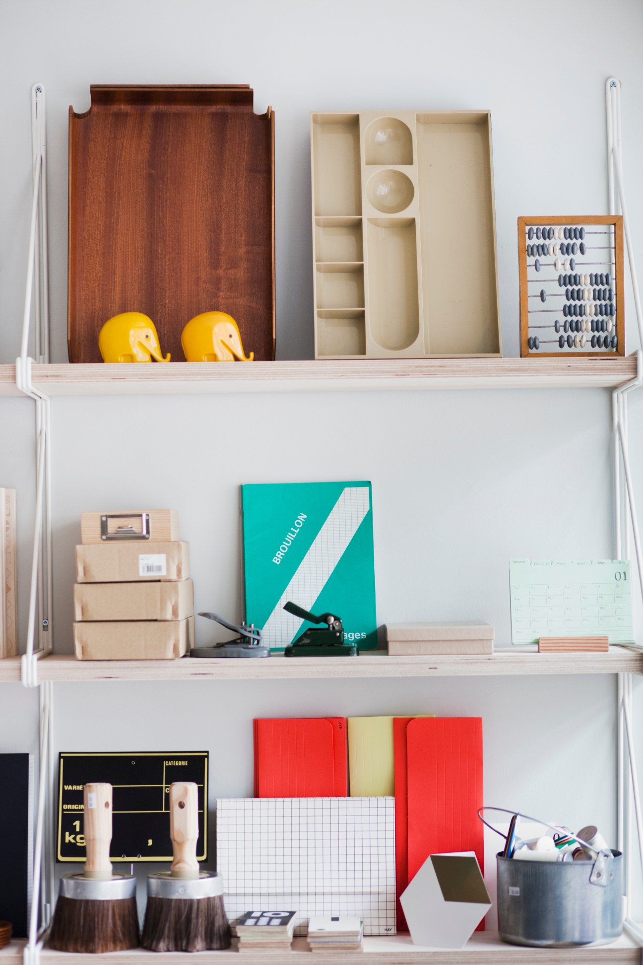 Shelving unit displaying stationery and design objects including wooden containers, yellow ceramic birds, notebooks, and office supplies.