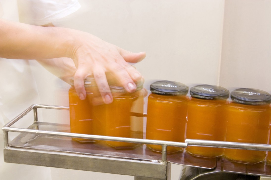 Hand reaching toward jars of amber-colored preserves with Staud's labels in a metal display tray.