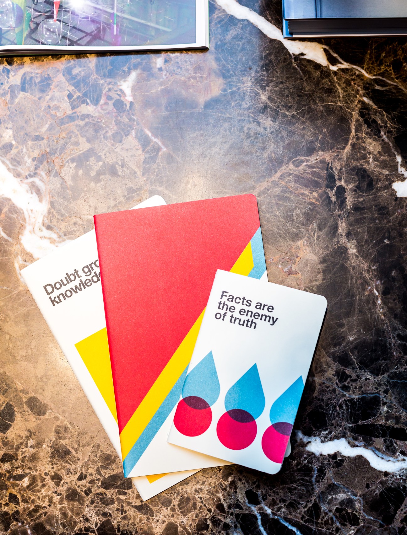 Two colorful books with geometric designs displayed on a marble surface, including titles "Doubt grows knowledge" and "Facts are the enemy of truth".