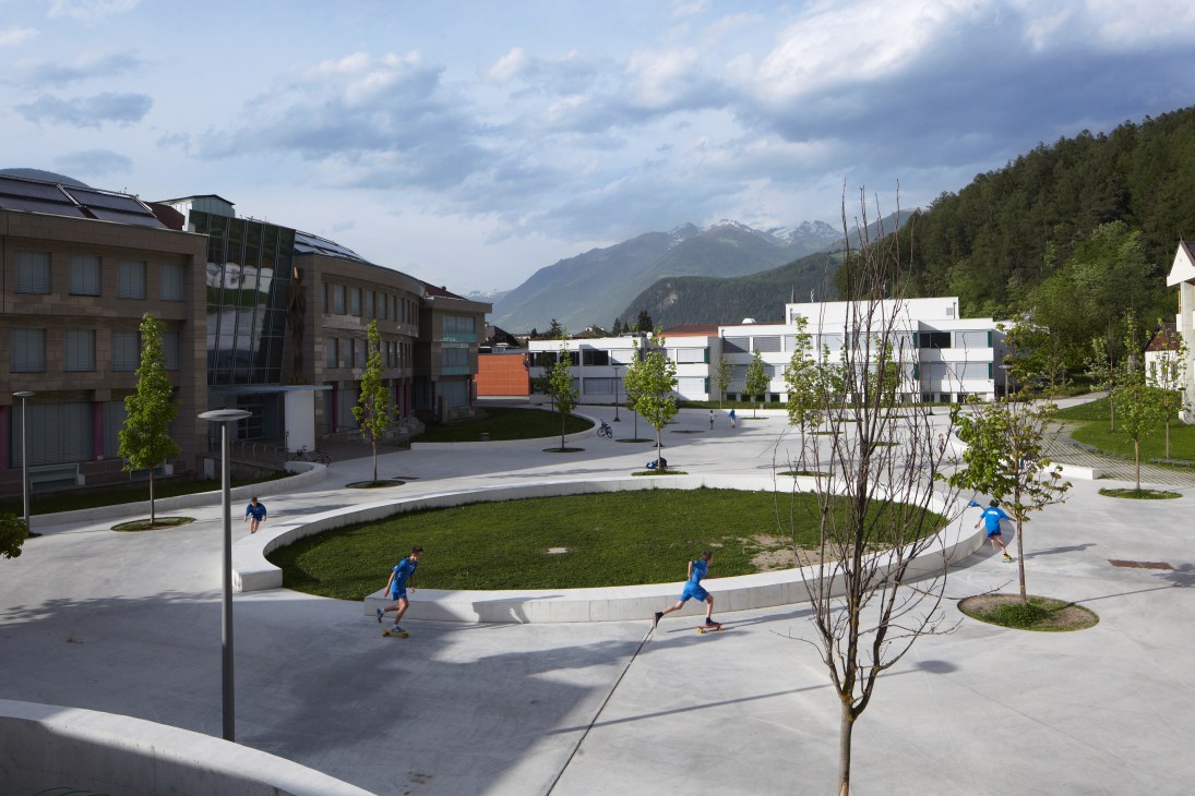 Children in blue shirts playing and running in a modern school playground in Brunico with green lawn, paved areas, young trees, and alpine mountains in the background.