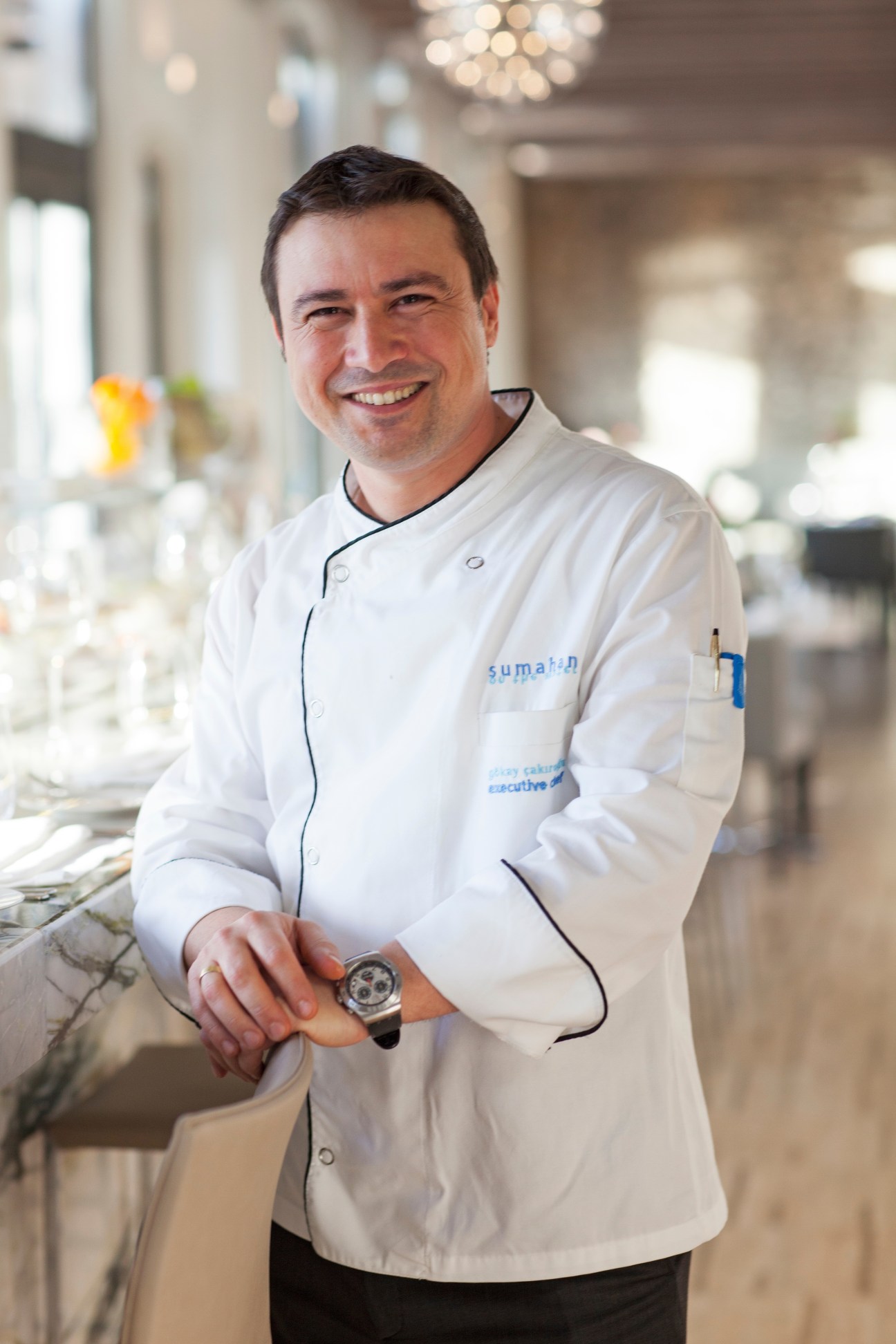 Chef Gokay Cakiroglu wearing white chef's coat smiles in an elegant dining room.