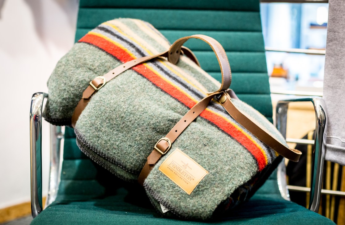 Green wool blanket with colorful stripes and leather handles displayed on a green bench.