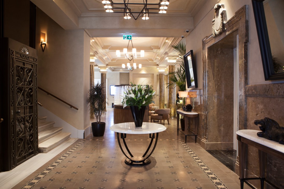 Elegant hotel entrance hallway with ornate marble flooring, decorative doors, and central round table with flowers.