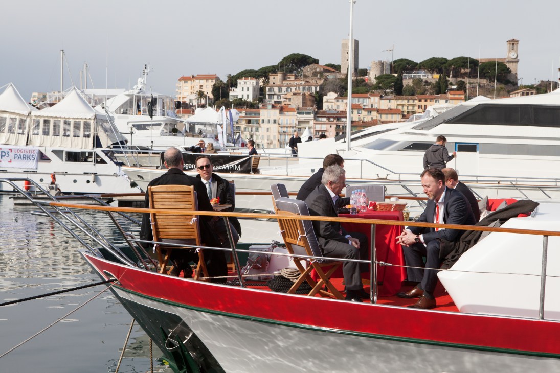 Business professionals in suits conduct meetings on a red and white yacht moored in Cannes marina with hillside buildings in background.