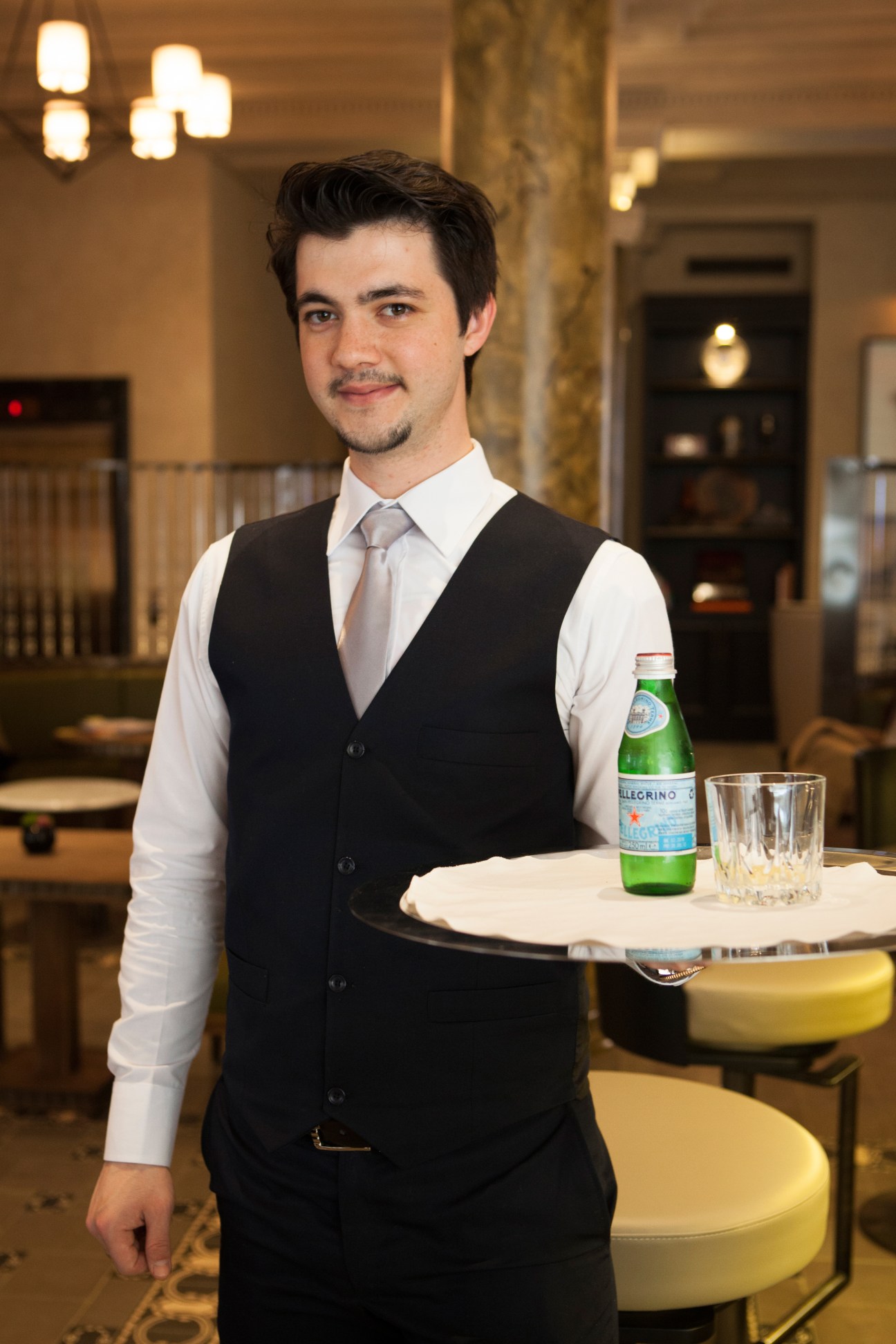 A male waiter in formal attire holding a tray with a bottle of water and glass in an elegant hotel lobby.