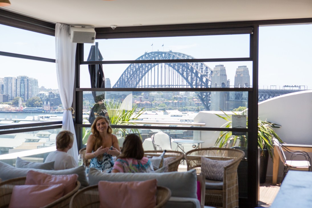 People relaxing in a rooftop lounge with floor-to-ceiling windows framing Sydney Harbour Bridge and city skyline views.