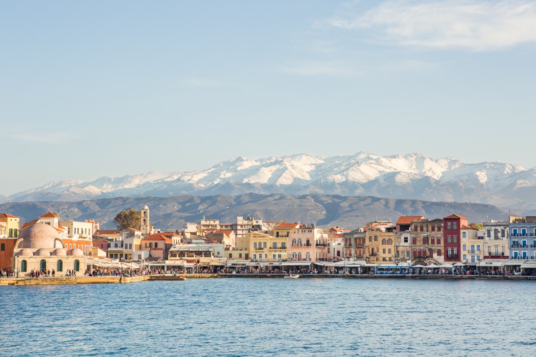 Colorful waterfront buildings of Chania port with snow-capped mountains in the distance across the Aegean Sea.