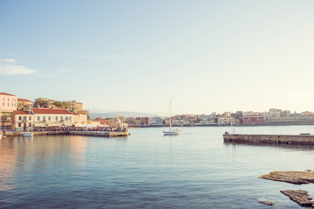 Tourists gathering at a waterfront promenade in Chania with colorful Venetian buildings and sailboats in the harbor.