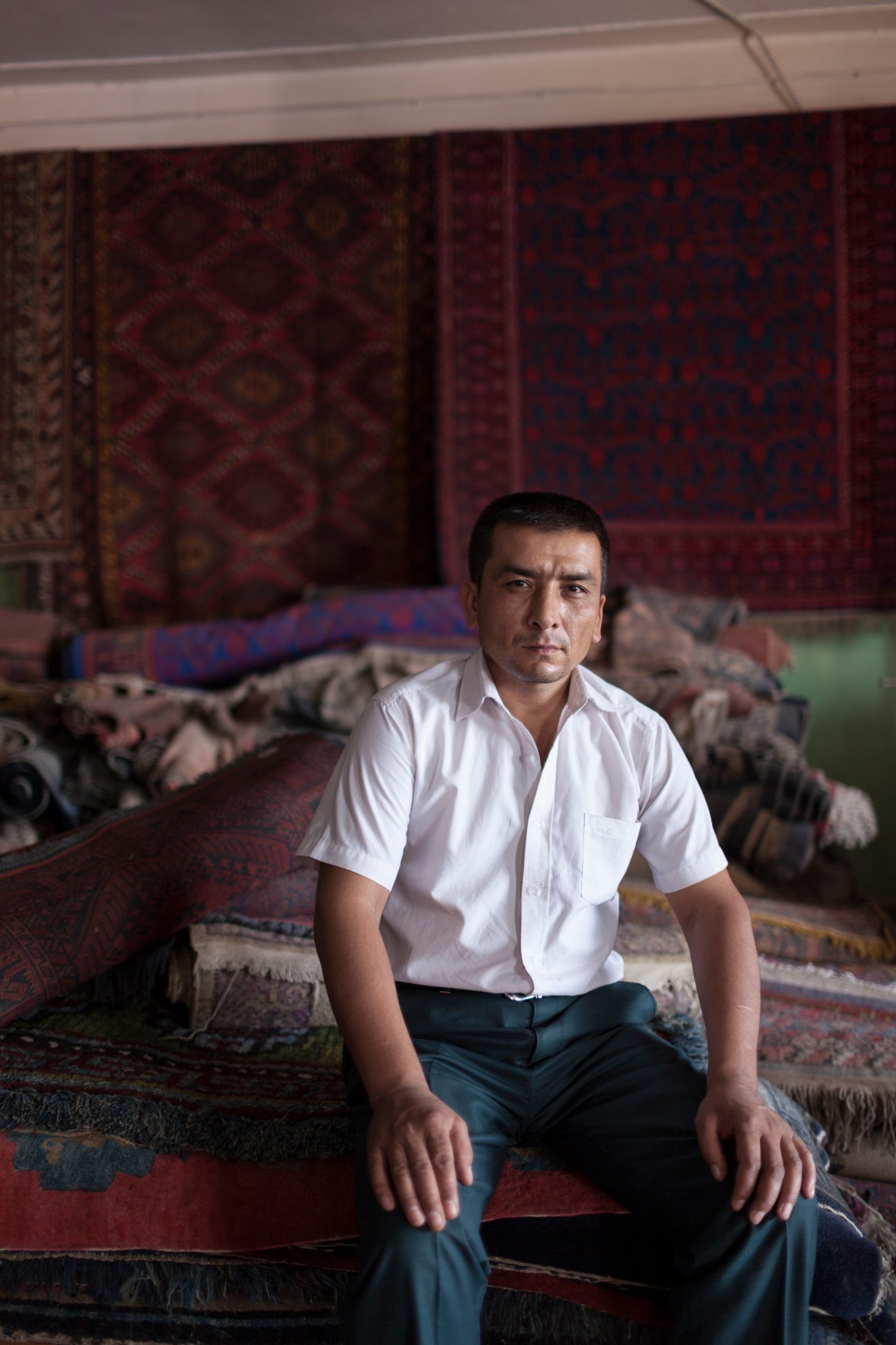 Muhammad Ali, a carpet dealer, sits on traditional rugs in his shop in Kashgar with ornate carpets hanging behind him.